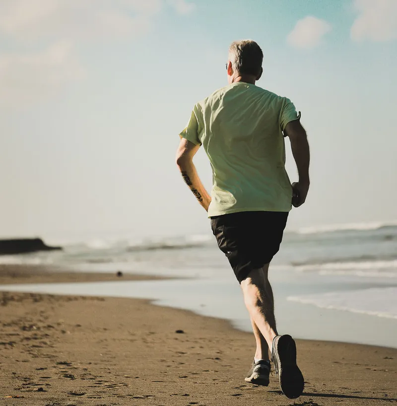 Man in green shirt jogging along a sandy beach shoreline next to calm waves.